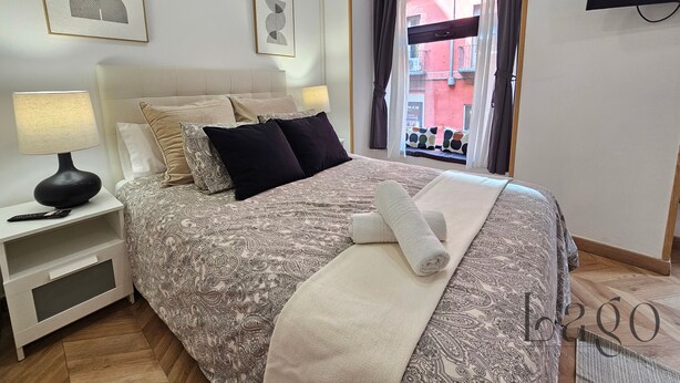 Ein ordentlich eingerichtetes Schlafzimmer mit Bett, Nachttisch, Lampe und einem Kunstwerk an der Wand.