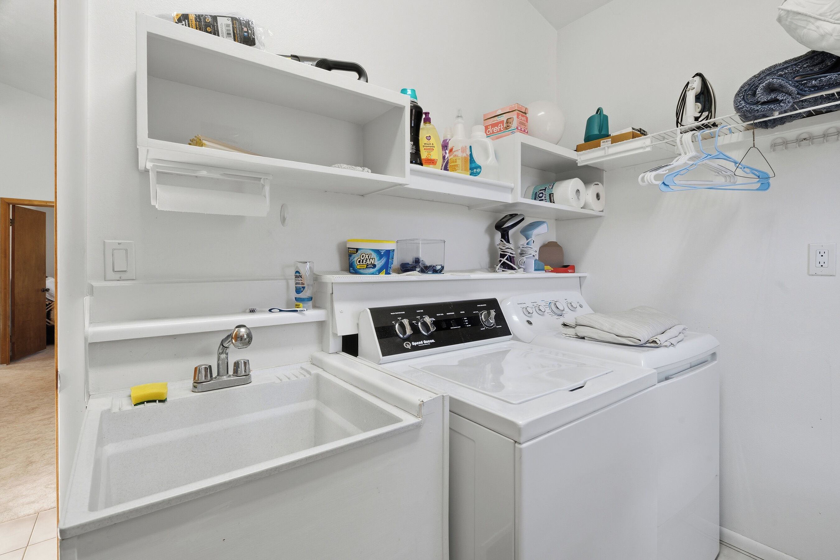 A laundry area with a washing machine, a dryer, and a sink. There are shelves above the sink with various items, including cleaning supplies and a box of toilet paper.