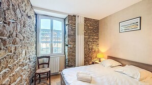 Une chambre avec un mur en pierre, une chaise en bois, un lit avec du linge de lit blanc, une lampe et une fenêtre ornée de rideaux à motifs.