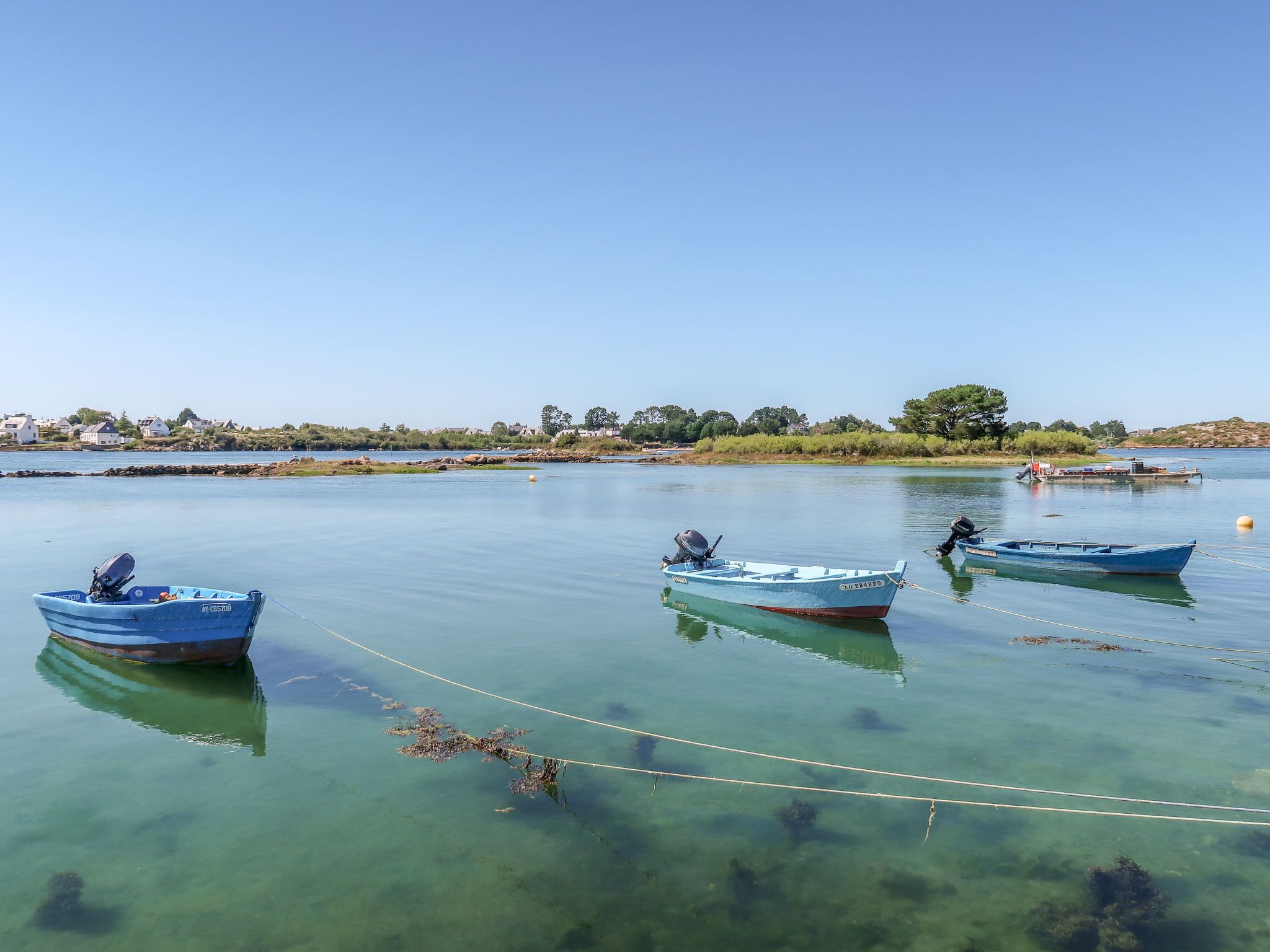 Een rustig meer met drie kleine boten afgemeerd in helder water, omgeven door een kalme oever met huizen en begroeiing.
