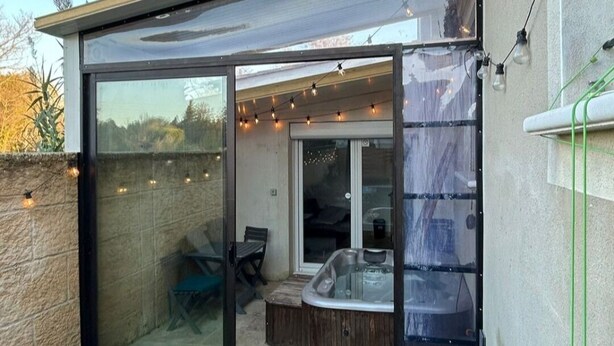 Une terrasse extérieure moderne, dotée d’une véranda en verre, d’un jacuzzi et de guirlandes lumineuses.