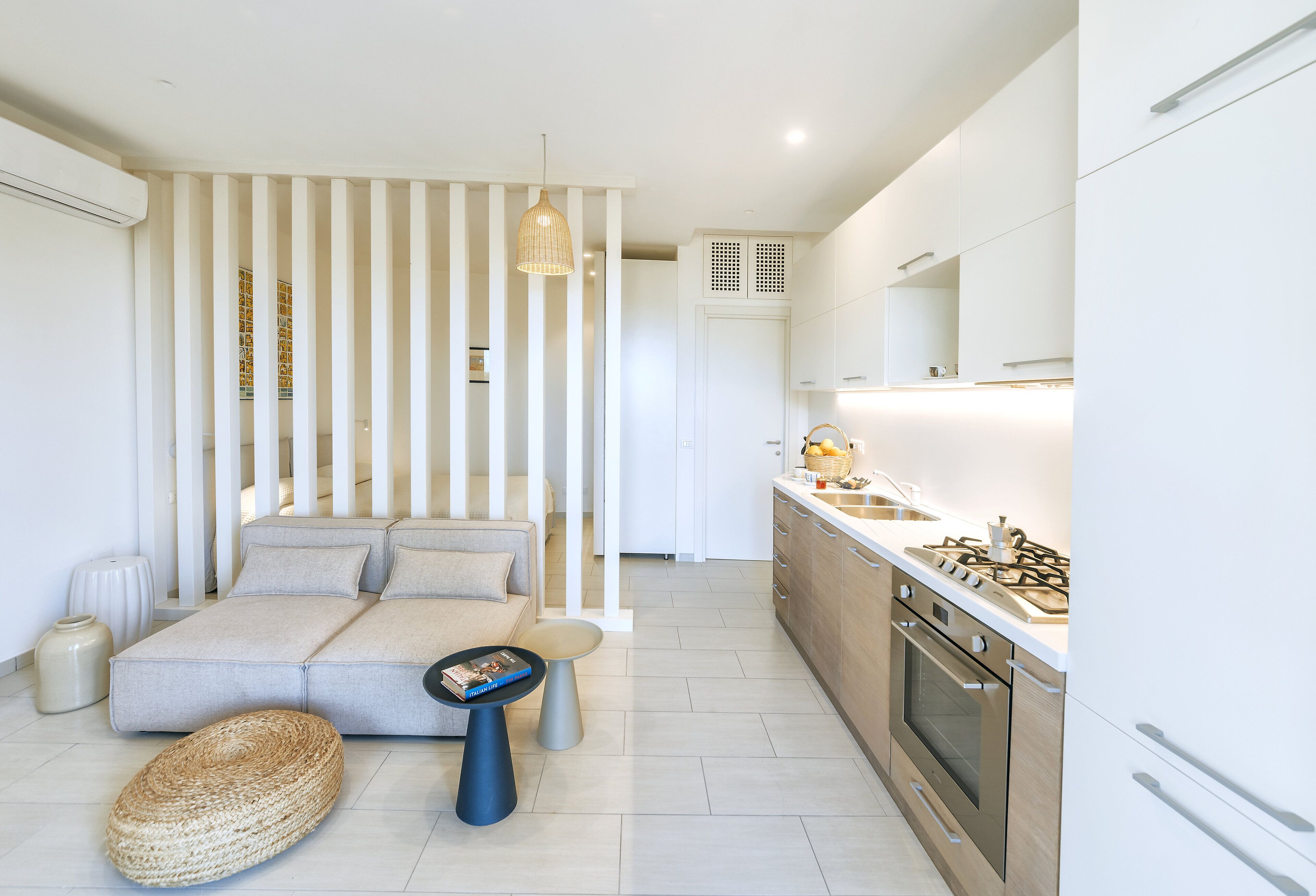 Une cuisine moderne équipée d’une cuisinière à gaz, d’un four et de meubles blancs. On y trouve un canapé avec des coussins et une petite table d’appoint sur laquelle repose un livre.