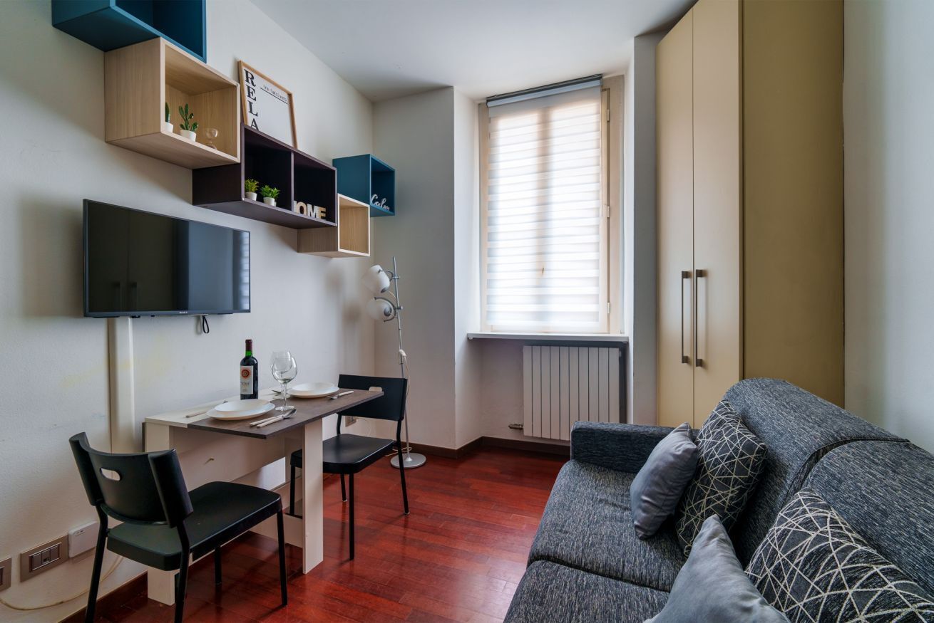 Un ambiente compatto con divano, tavolo da pranzo e sedie, televisione a parete e scaffali a muro con piante.
