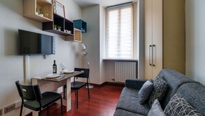 A compact living space with a sofa, a dining table with chairs, a TV mounted on the wall, and built-in shelving with plants. - Cadorna's Nest - Cozy and Central (Milano)