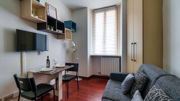Un ambiente compatto con divano, tavolo da pranzo e sedie, televisione a parete e scaffali a muro con piante.