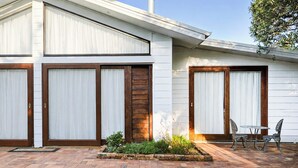 Une maison moderne avec deux grandes fenêtres, un chemin en briques et un petit jardin agrémenté de chaises.