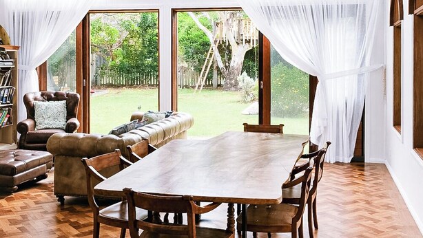 Une vaste pièce aménagée comme une tente, avec une grande table et des chaises en bois, des rideaux blancs et de grandes fenêtres donnant sur un jardin.