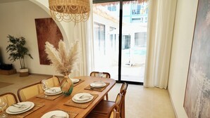 A dining area with a long wooden table set for four, surrounded by wicker chairs, with a large window offering a view of a pool and building. - MaShang Holiday Villas - Luxury 5BR Private Pool Villa in Jomtien (Muang Pattaya)