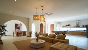 A spacious living area with a round coffee table, a sofa, and a dining table with chairs. There are hanging pendant lights and a kitchen in the background. - MaShang Holiday Villas - Luxury 5BR Private Pool Villa in Jomtien (Muang Pattaya)