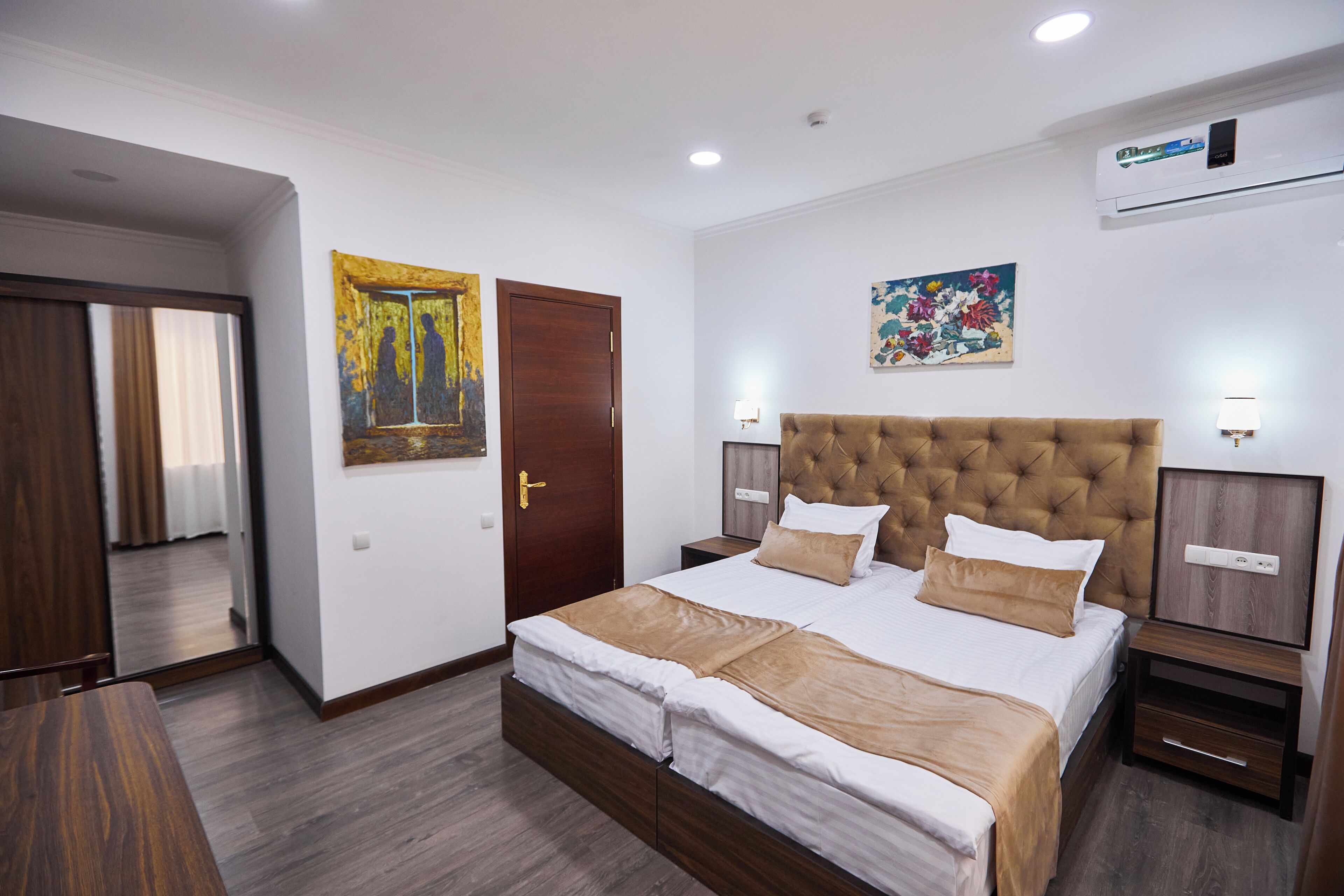 A hotel room with a large bed, a wooden door, a painting, a nightstand, and a wall-mounted air conditioner.