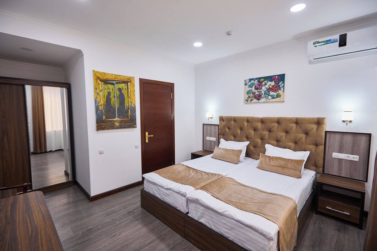 A hotel room with a large bed, a wooden door, a painting, a nightstand, and a wall-mounted air conditioner.