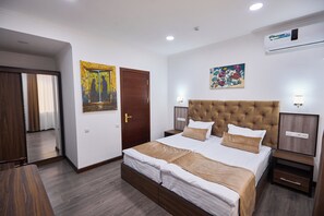 A hotel room with a large bed, a wooden door, a painting, a nightstand, and a wall-mounted air conditioner. - Art Grand  (Tashkent)