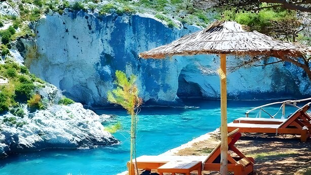 Una playa con sillones de madera y una sombrilla de paja, con vista a un mar azul cristalino y acantilados rocosos.