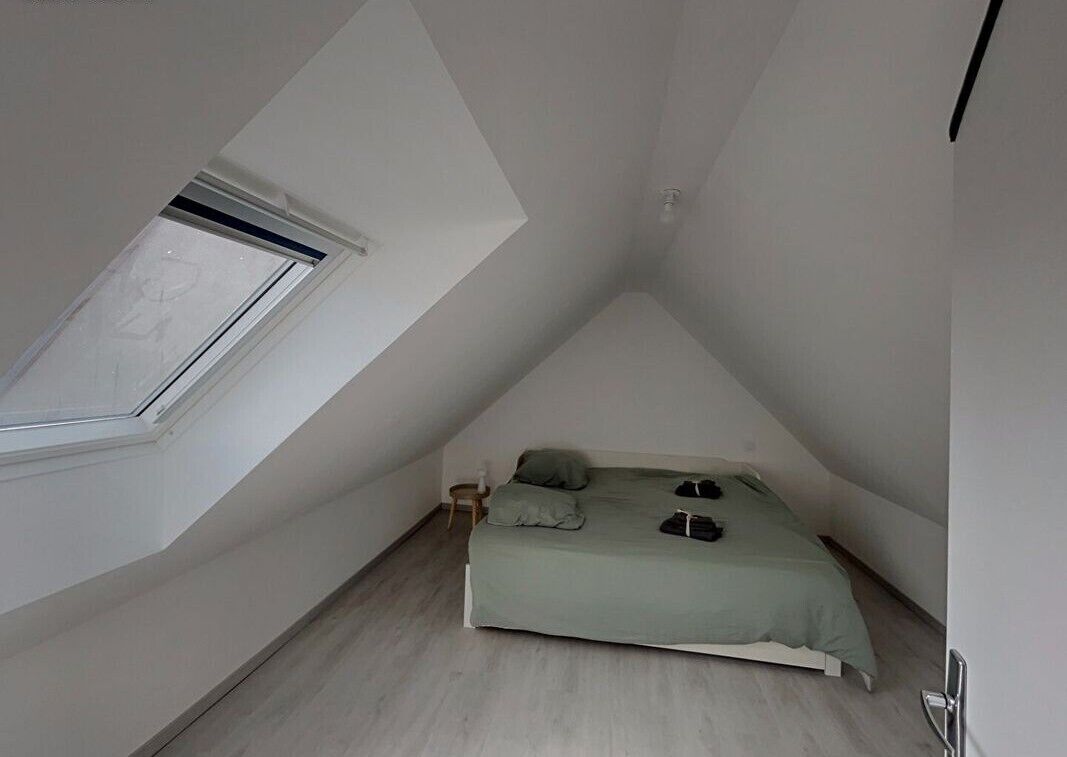 Une petite pièce triangulaire avec un plafond en pente et un puits de lumière. On y trouve un lit recouvert d’une couverture verte, deux oreillers et un petit tabouret en bois.