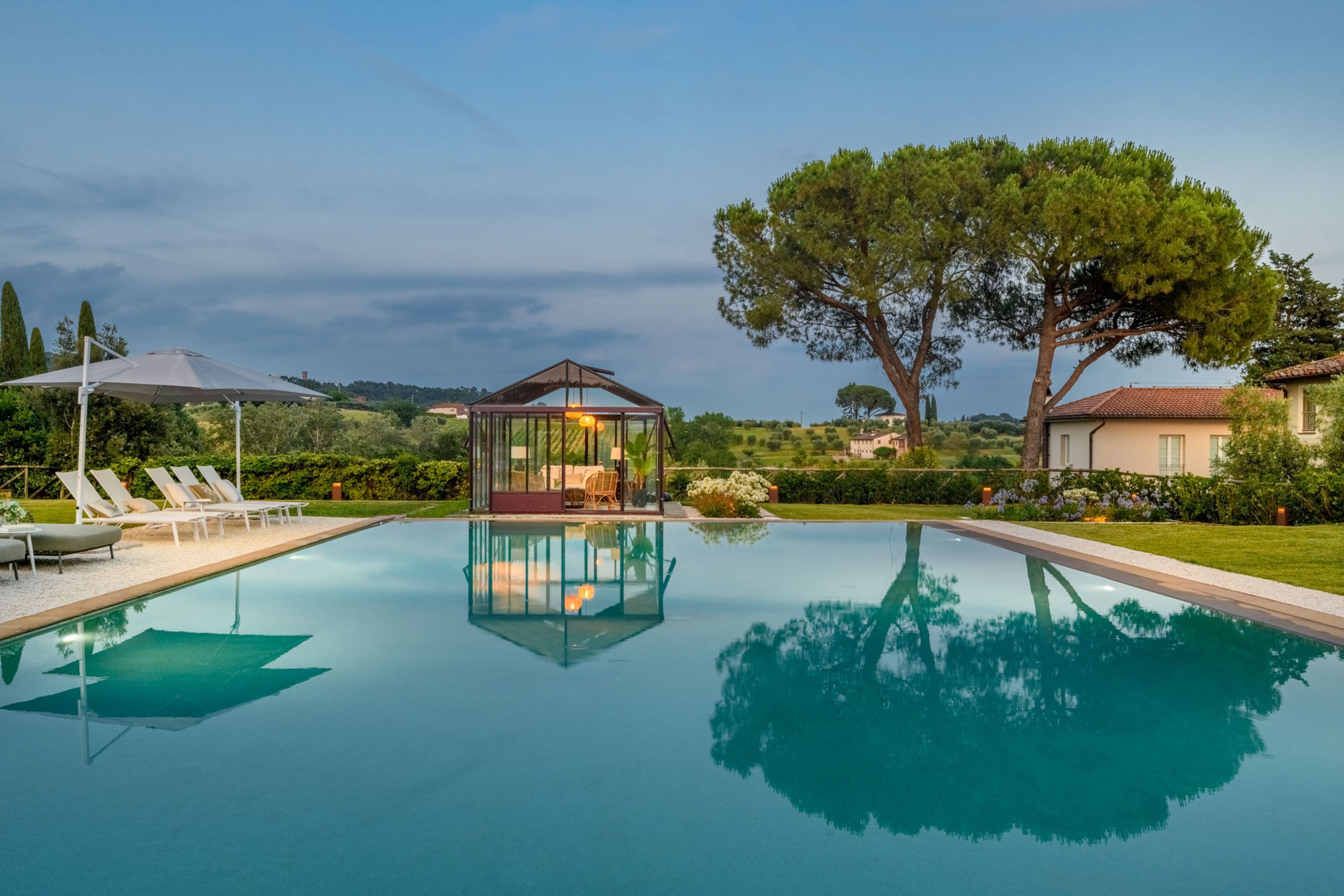 A swimming pool with a glass-enclosed gazebo, lounge chairs, and a large tree.