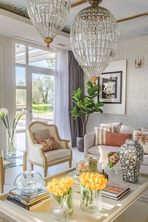 A well-lit living room with a white brick wall, a large window, and a chandelier. There is a sofa, a chair, and a coffee table with books and vases. - Corte Benedetto in Montecarlo (Capannori)