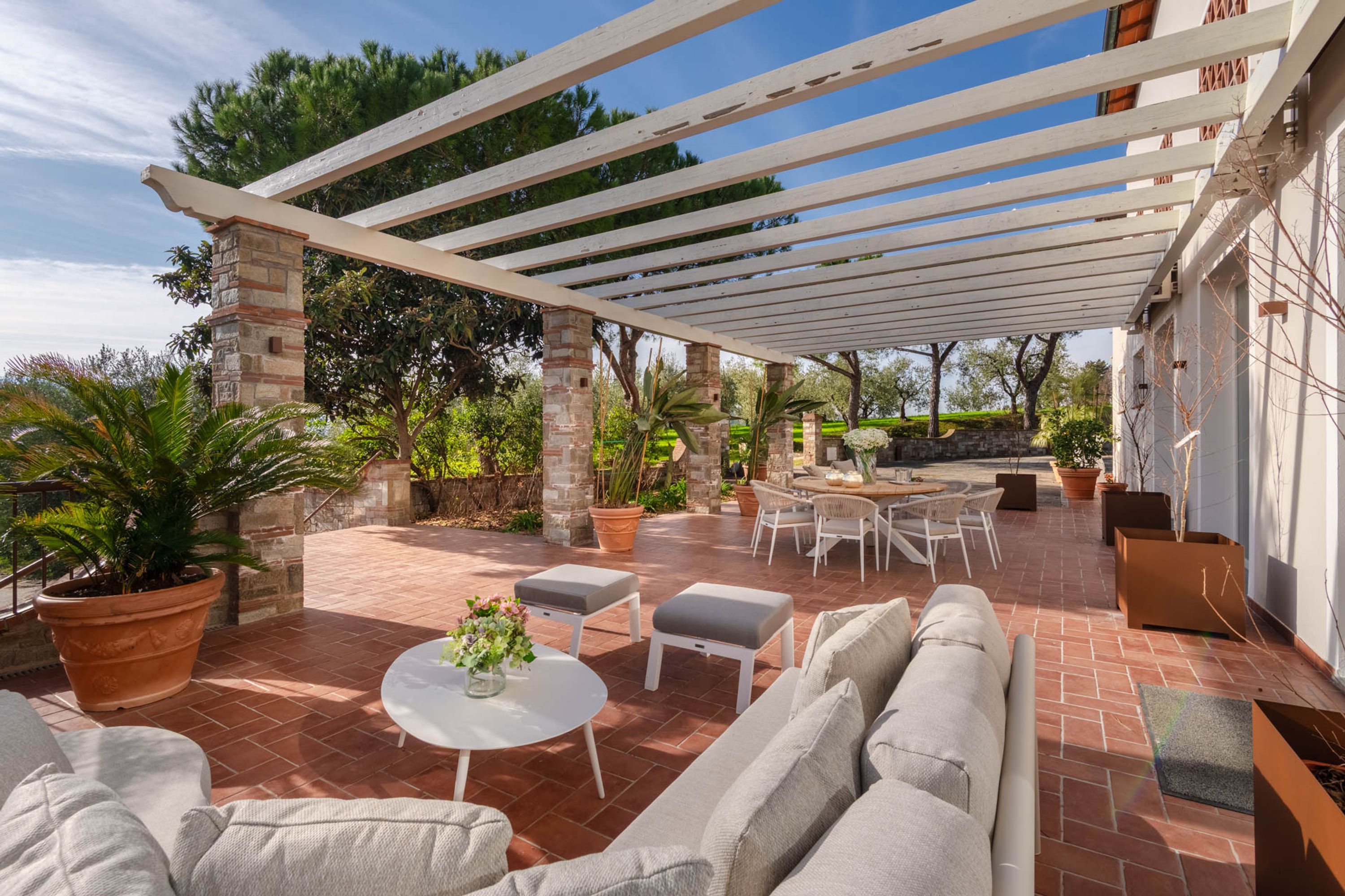 Un patio techado con una pérgola blanca, mobiliario exterior y plantas en macetas.