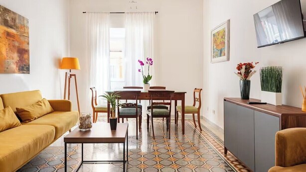 A modern living room with a sofa, coffee table, dining table, chairs, and a TV mounted on the wall. - Rhome Elegant Apartment (Roma)