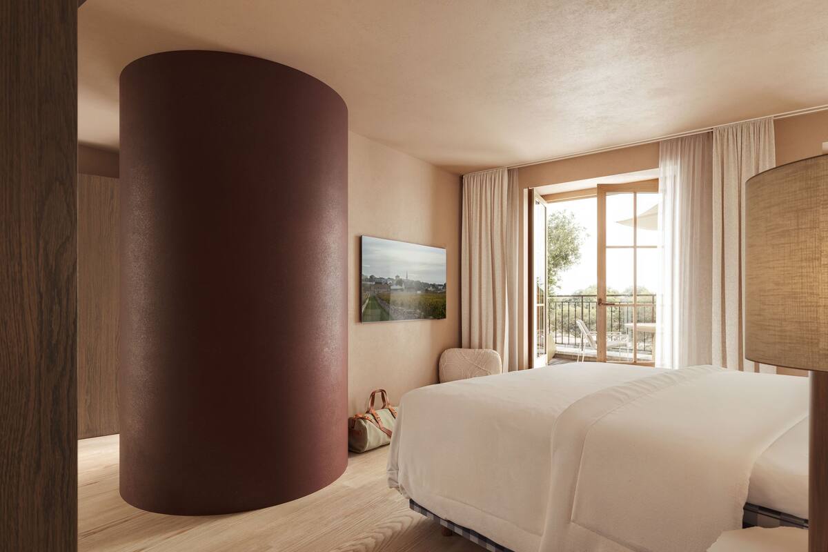 A hotel room with a large bed, a window with curtains, a painting on the wall, and a wooden pillar.