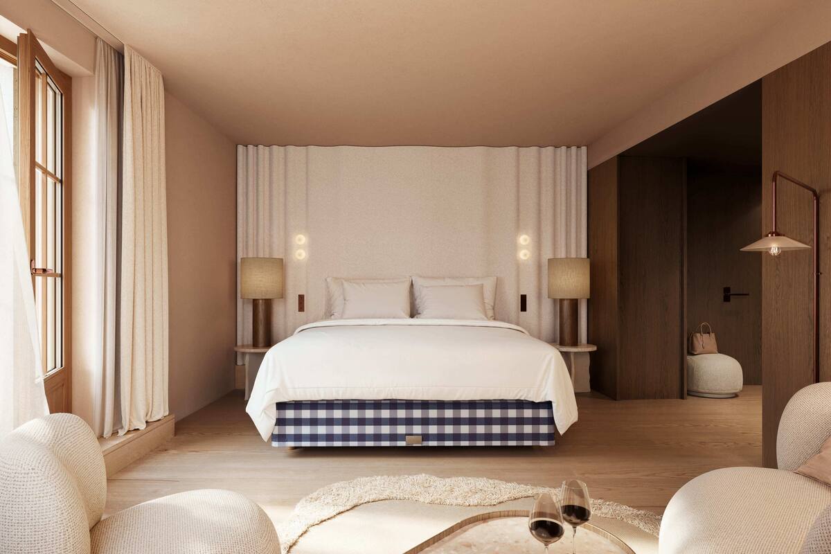 A bedroom with a large bed, two bedside lamps, a nightstand, a window with curtains, and a wooden floor.