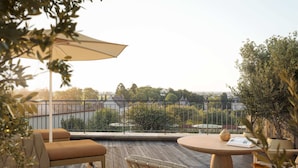 Una terraza de madera con mobiliario exterior, una mesa redonda y vistas a un paisaje urbano.