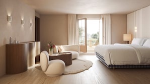 Una habitación de hotel moderna con una cama grande, una zona de estar con una mesita de café, un escritorio con una computadora y vistas al exterior a través de un ventanal amplio.