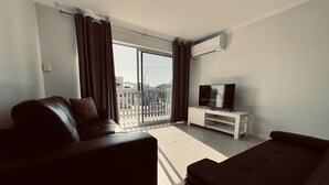 A modern living room with a sofa, a TV on a white stand, and a balcony with a view of the street. - Great Location Close to the City and Strand - Great Location Close to the City and Strand (North Ward)