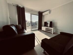 A modern living room with a sofa, a TV on a white stand, and a balcony with a view of the street. - Great Location Close to the City and Strand - Great Location Close to the City and Strand (North Ward)