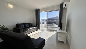 A modern living room with a black leather sofa, a white side table, and a large sliding glass door leading to a balcony with a view of the street. - Great Location Close to the City and Strand - Great Location Close to the City and Strand (North Ward)