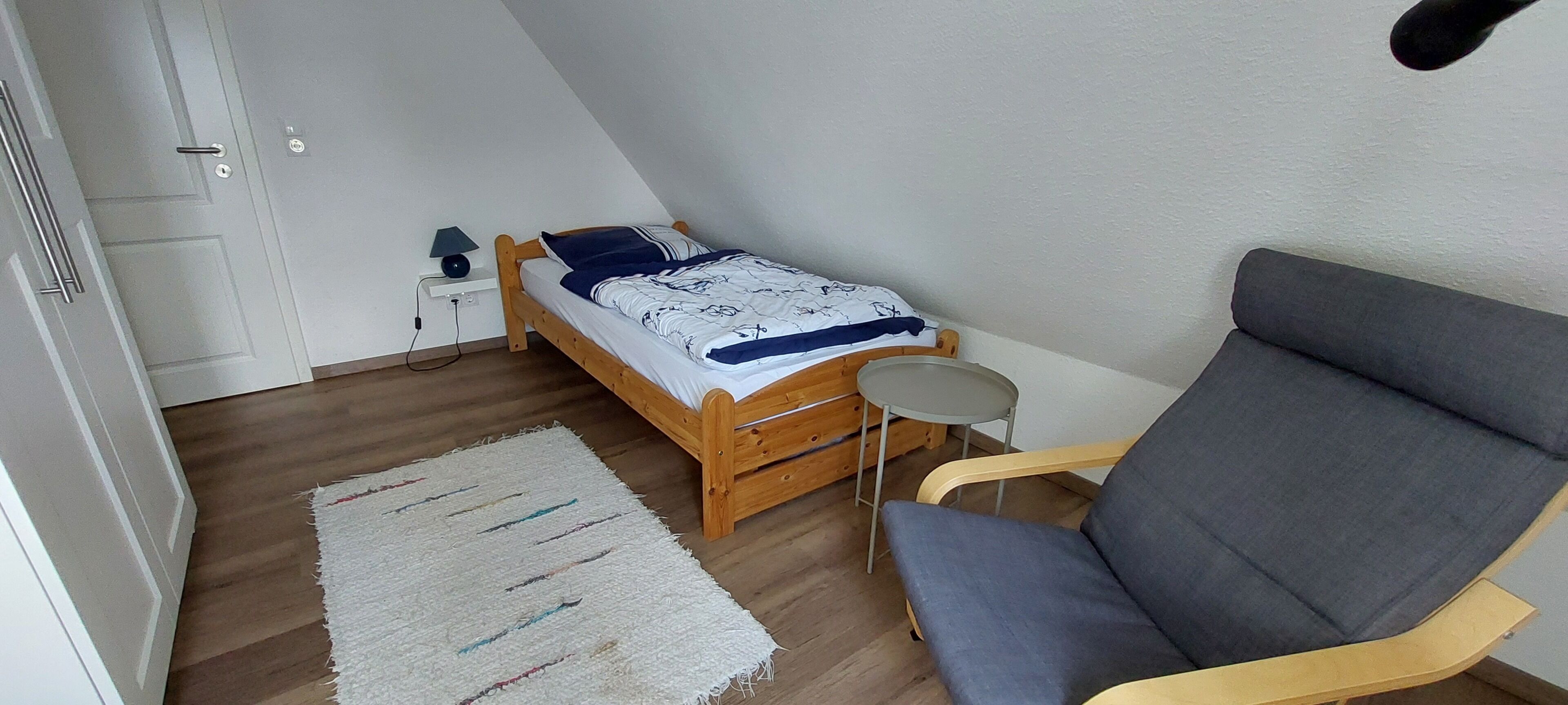 A bedroom with a bed, a chair, a small table, a lamp, and a door.