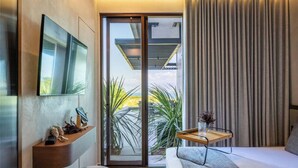 Et moderne hotelværelse med en stor seng, et fladskærms-tv monteret på væggen, et lille trænatbord og en skydedør i glas, der fører ud til en balkon med planter.