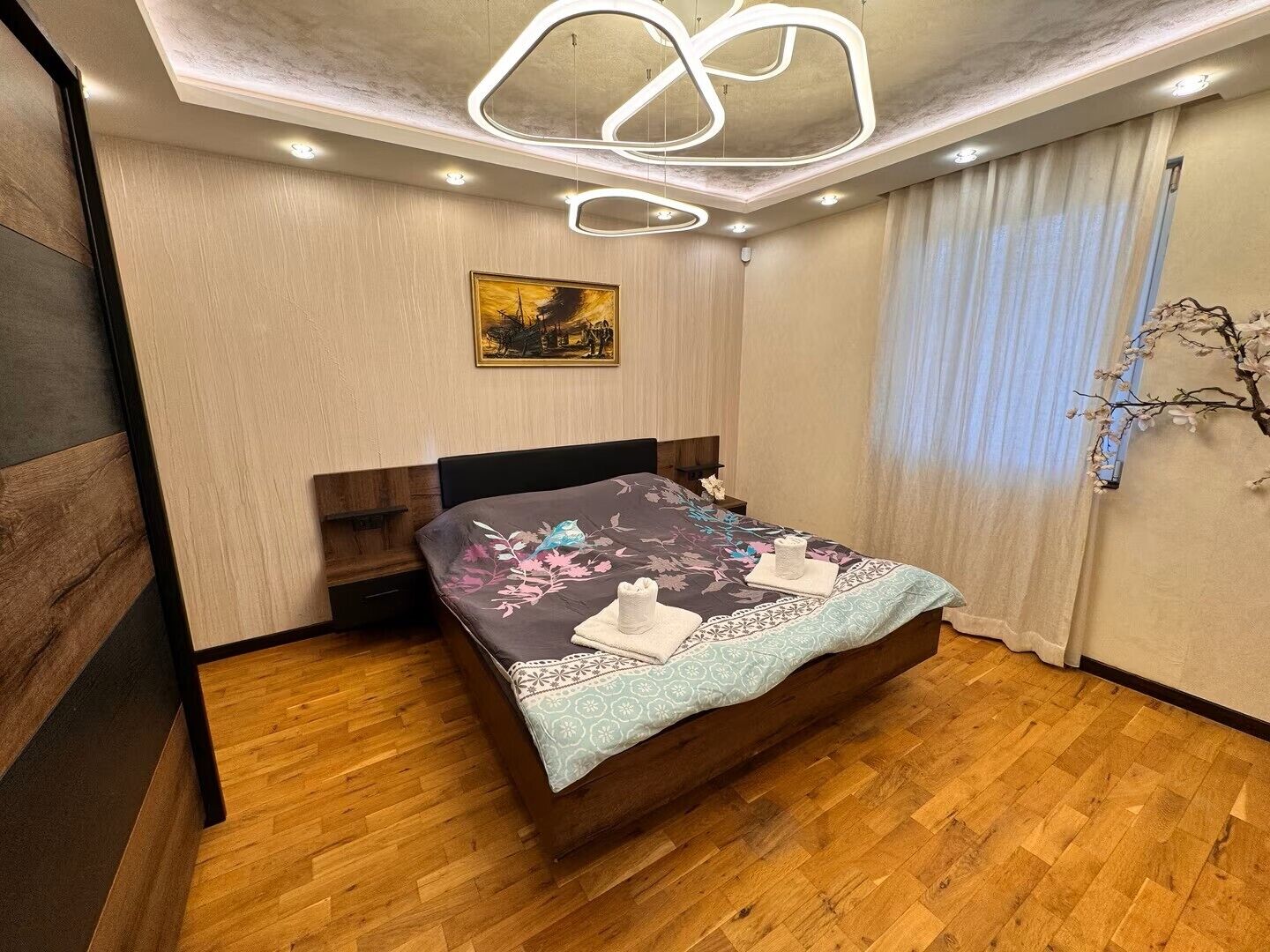 Una camera da letto moderna con pavimento in legno, un letto con biancheria da letto floreale, un quadro appeso al muro e un lampadario dal design originale.