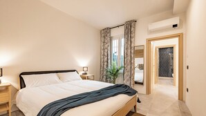 Quarto moderno com uma cama grande, mesas de cabeceira, uma planta em vaso e vista para o banheiro através de uma porta de vidro.