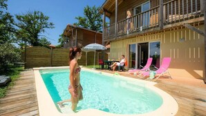 En træterrasse med swimmingpool, en overdækket terrasse og en toetagers bygning med balkon.