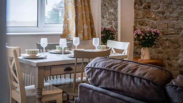 A dining area with a stone wall, a window with a curtain, and a table set for two with wine glasses and bowls. - Tide & Tee - 2 Bedroom Apartment - Penally - Tenby (Tenby)