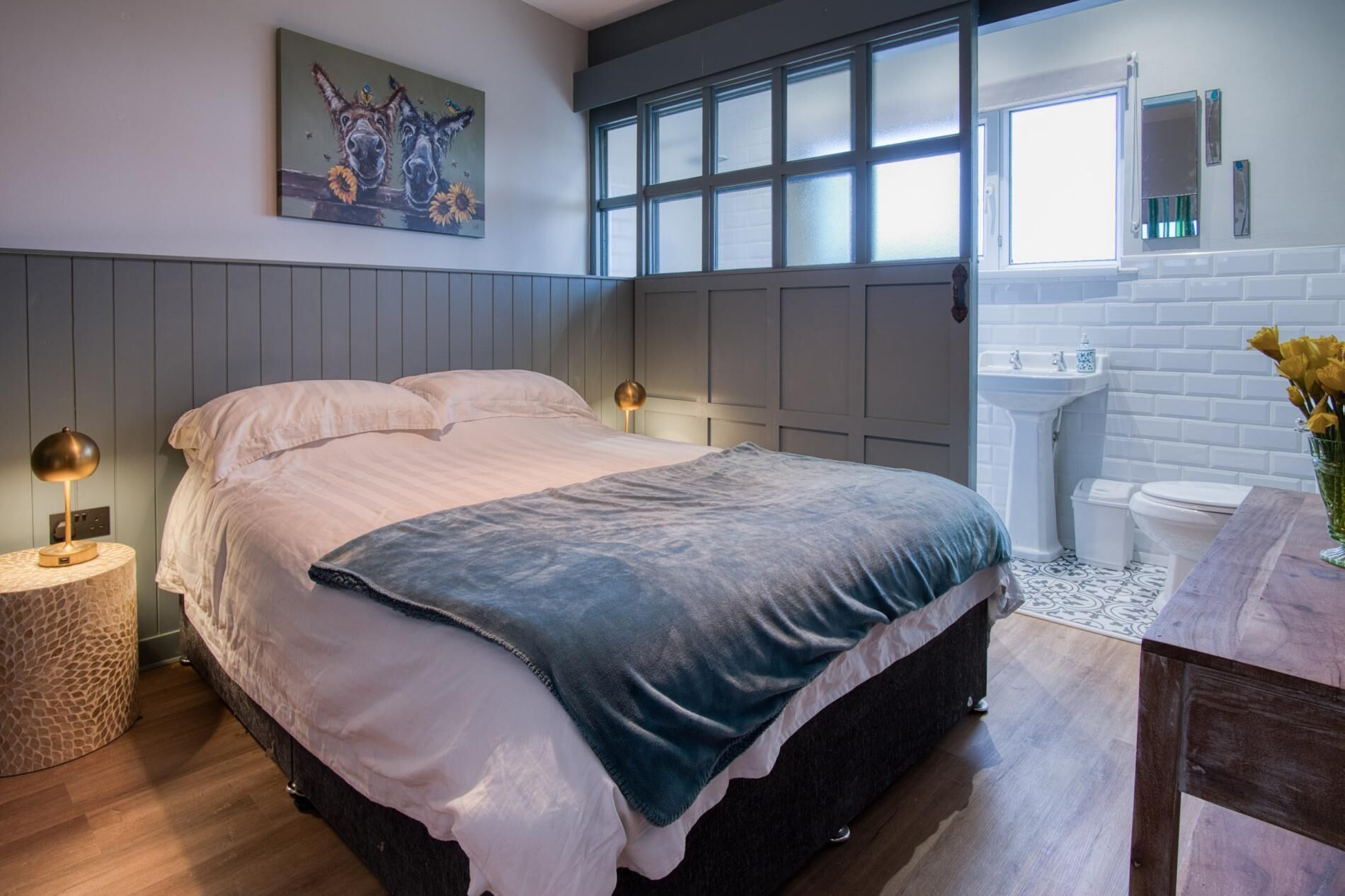 A modern bedroom with a large bed, bedside lamps, a painting, a window, and a bathroom with a sink and toilet.