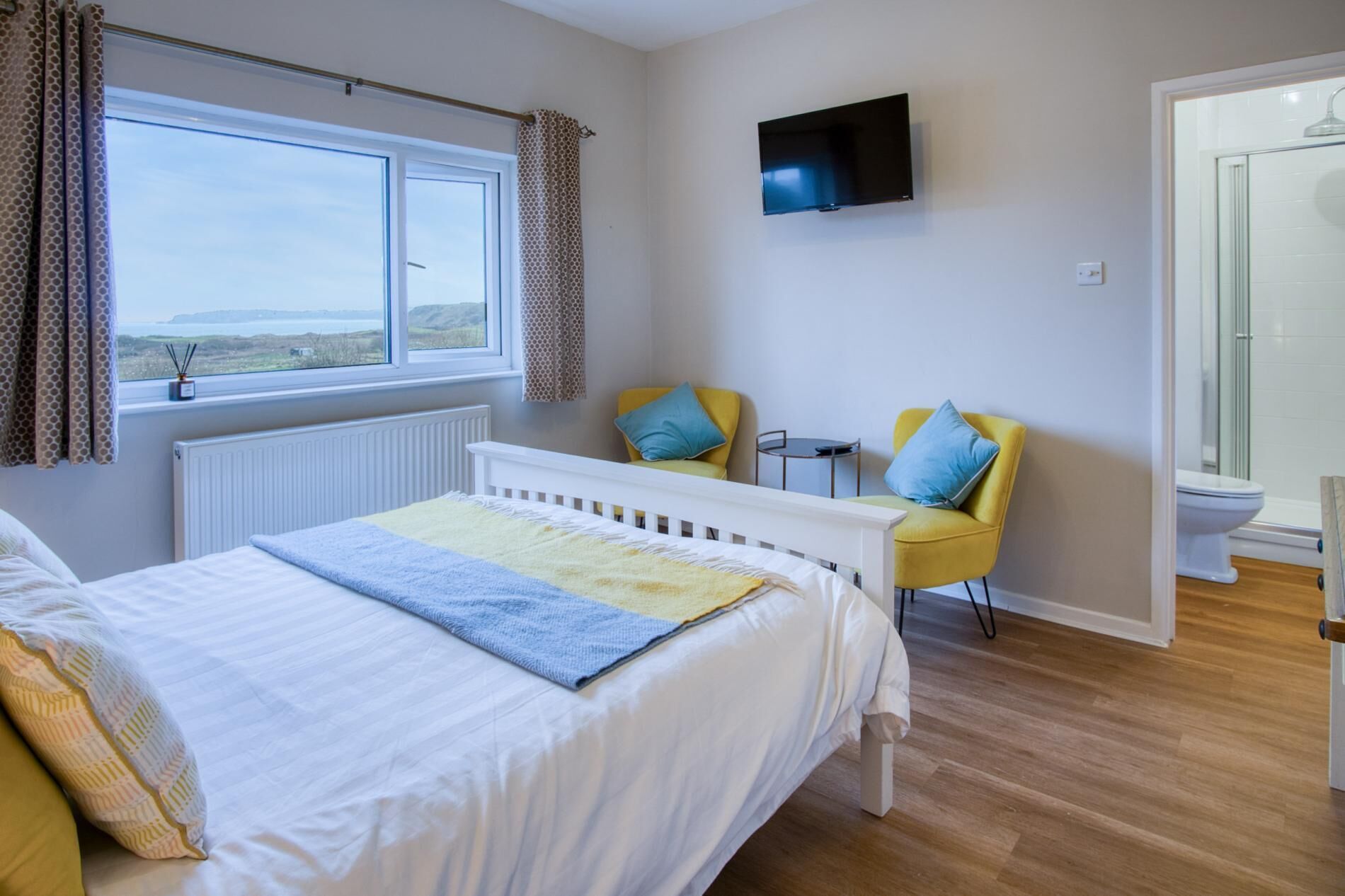 A bedroom with a bed, a TV mounted on the wall, a small table, two chairs, and a view of the outdoors through a window.