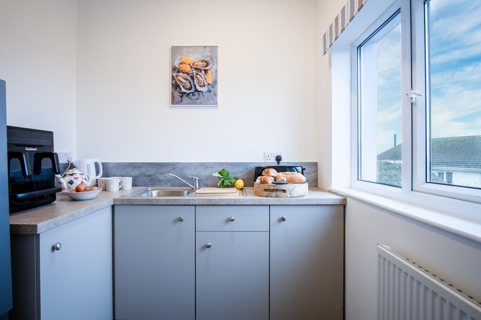 Et moderne køkken med lysegrå skabe, kaffemaskine, vask og et vindue med udsigt til en bygning.