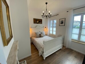 A bedroom with a white bed, a chandelier, and a window with a view.