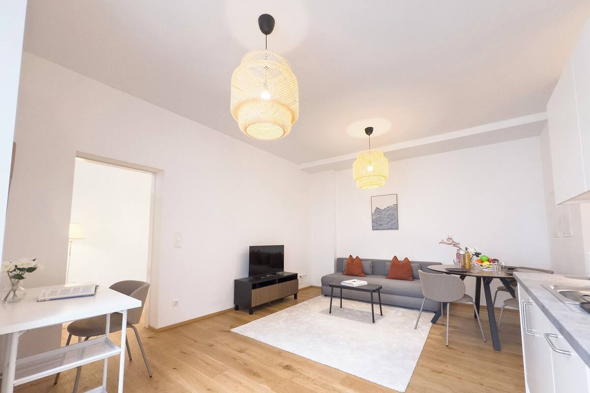 Un salon moderne avec un canapé, une table basse et un téléviseur. Il y a également un coin repas avec une table et des chaises. La pièce est dotée d’un parquet et de murs blancs.