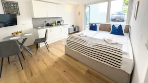 A modern hotel room with a bed, a small kitchenette, a dining area, and a large window offering a view of the street. - Hillside Living Vienna (Vienna)