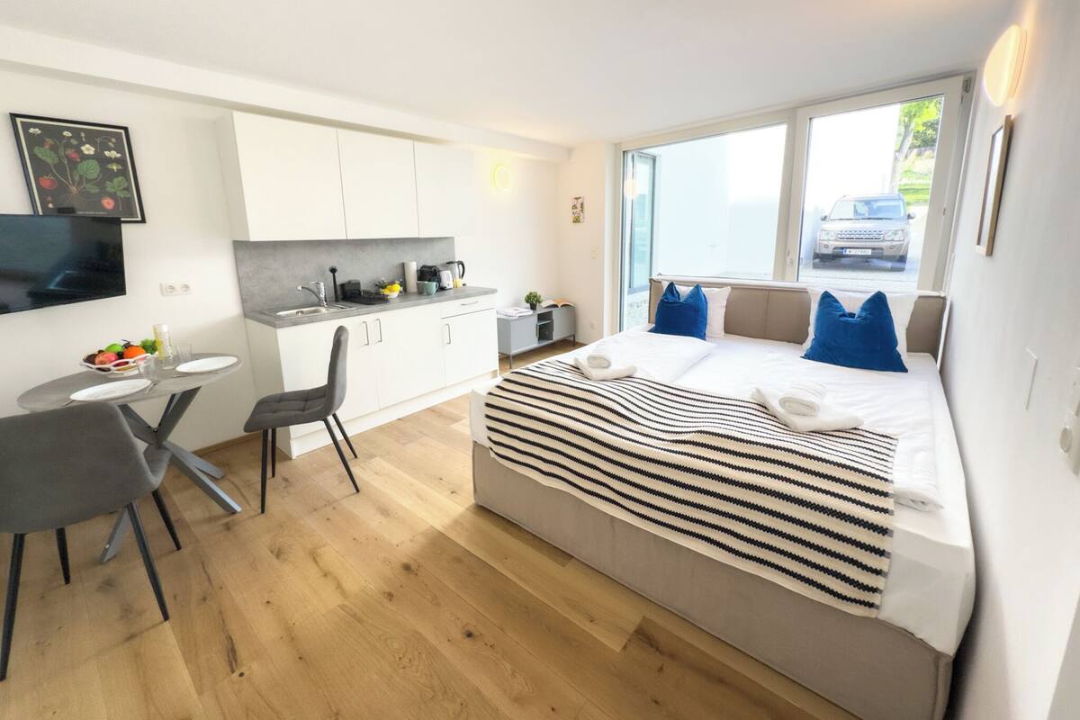 Une chambre d’hôtel moderne équipée d’un lit, d’une petite kitchenette, d’un coin repas et d’une grande fenêtre donnant sur la rue.