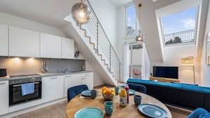 A modern kitchen with white cabinets, a built-in oven, and a dining area with a wooden table and blue chairs. - Hillside Living Vienna (Vienna)