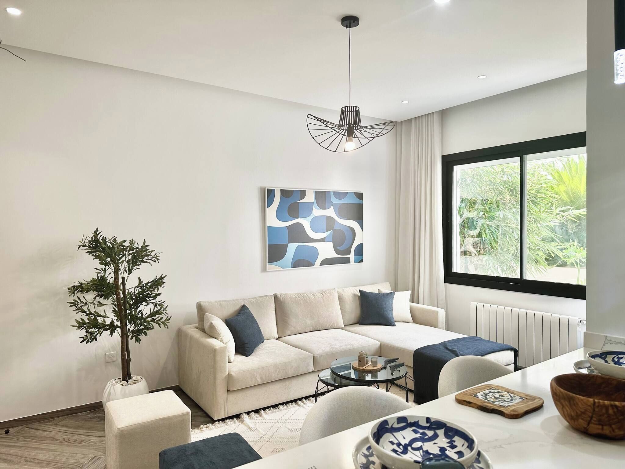 A modern living room with a sofa, coffee table, and a dining area with a table and chairs. There is a large window with a view of greenery outside.