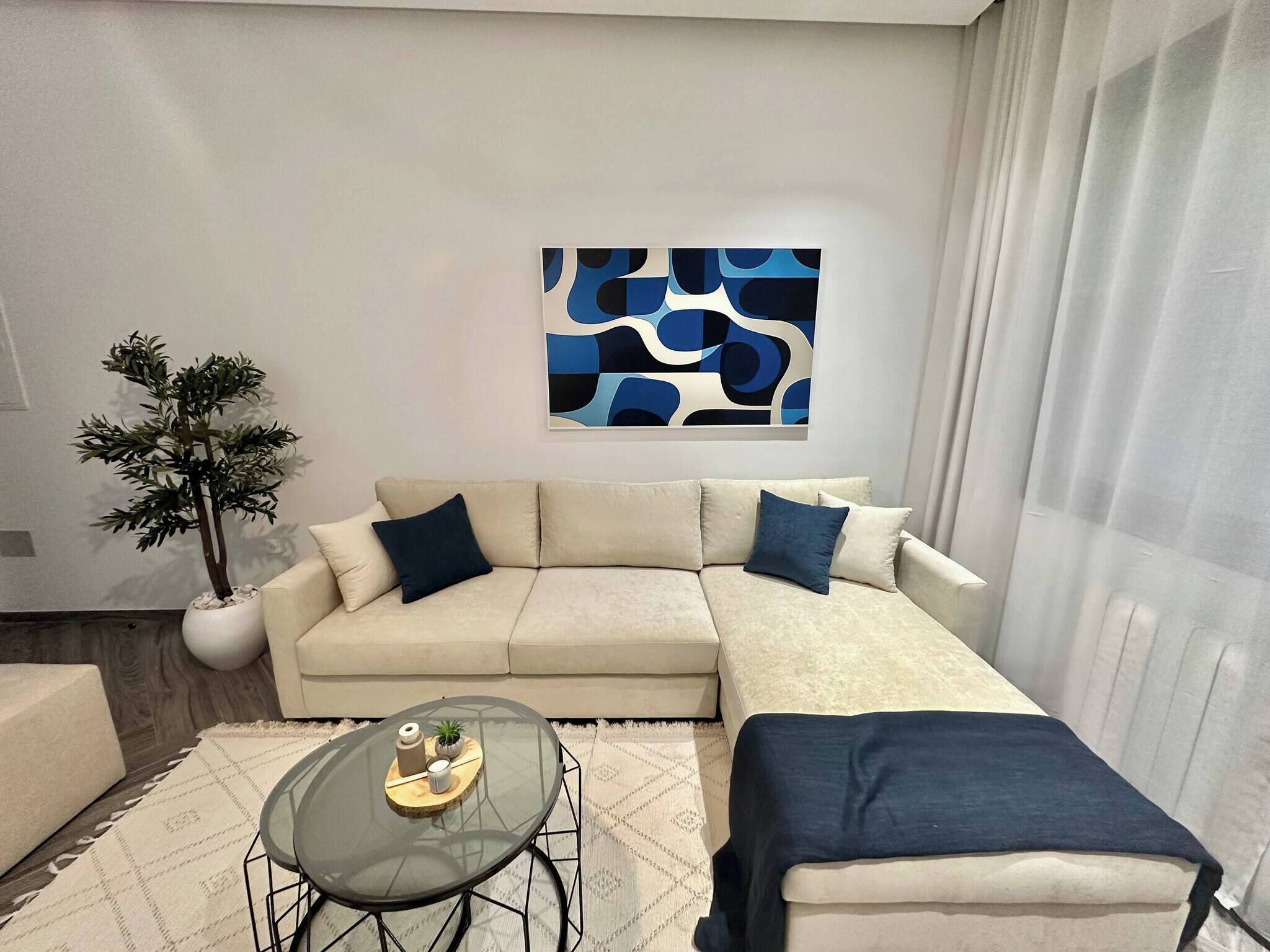 Un salon moderne avec un canapé d’angle beige, une table basse en verre, une plante en pot et un tableau abstrait bleu accroché au mur.