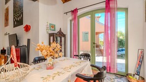 Un coin repas avec une table dressée pour deux personnes, un vase contenant des fleurs séchées, un buffet et une vue sur l’extérieur par une porte munie de rideaux.