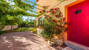 Un chemin pavé mène à un bâtiment doté d’une porte rouge, entouré de verdure et sous un ciel dégagé.