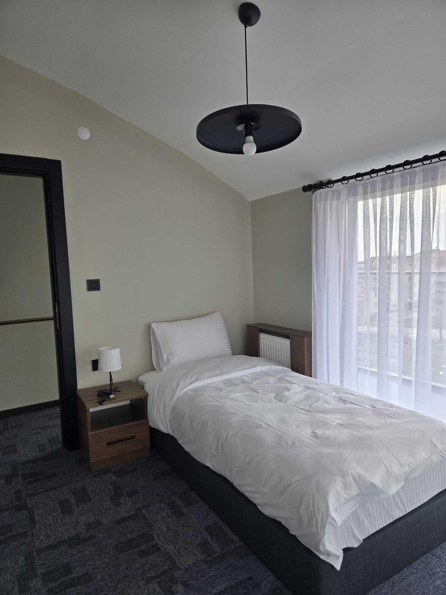 A hotel room with a bed, a bedside table with a lamp, a radiator, and a window with sheer curtains.