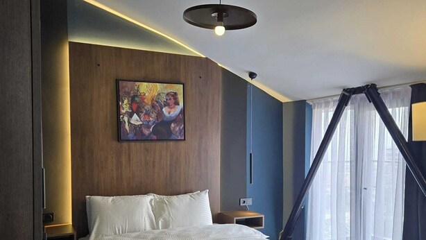 A modern hotel room with a large bed, a painting on the wall, a ceiling light, and a window with sheer curtains.