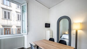 A hotel room with a wooden table, two chairs, a large mirror, a TV on the wall, and a window with a view of buildings. - Duomo collection suites B (Florencia)
